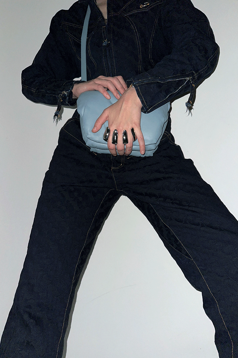 Person wearing a dark denim outfit holding a blue handbag against a white background