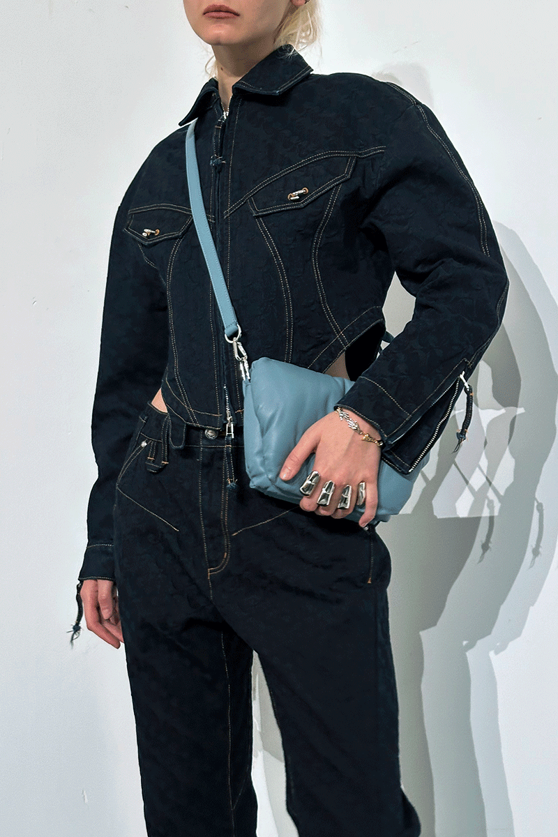 Person wearing a dark denim outfit holding a blue handbag against a white background